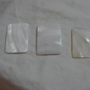 May include: Three white rectangular mother of pearl tiles with a swirl pattern. Each tile has five small holes.