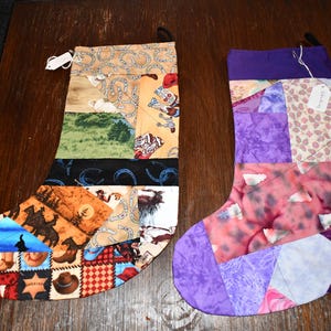 May include: Two handmade Christmas stockings. One is patchwork with cowboy-themed fabric, featuring horseshoes, boots, and hats. The other is a patchwork of purple, pink, and white fabrics. Both have a loop for hanging.