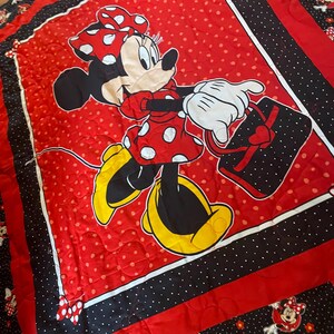 May include: Red and black Minnie Mouse quilt featuring a large central image of Minnie Mouse in a red polka dot dress and bow, carrying a black purse with a heart. The quilt has a red polka dot background, black and white polka dot borders, and Minnie Mouse character accents.