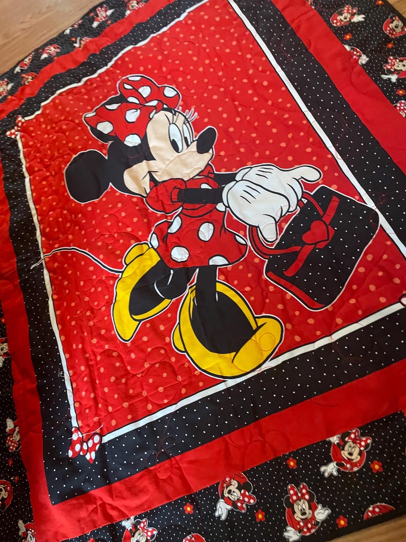 May include: Red and black Minnie Mouse quilt featuring a large central image of Minnie Mouse in a red polka dot dress and bow, carrying a black purse with a heart. The quilt has a red polka dot background, black and white polka dot borders, and Minnie Mouse character accents.