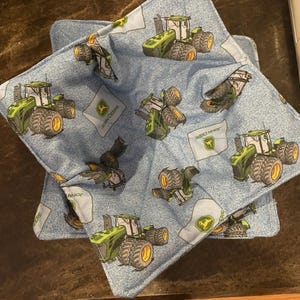 May include: Two denim-colored bowl cozies with a green and yellow tractor pattern. Each cozy features a square shape with a folded edge and a John Deere logo. The tractor design is repeated across the fabric.
