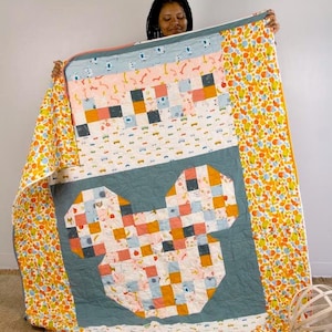 May include: A quilt with a Mickey Mouse design made from colorful patchwork fabric. The quilt is blue, pink, orange, and white. The quilt is folded in half and held by a person.