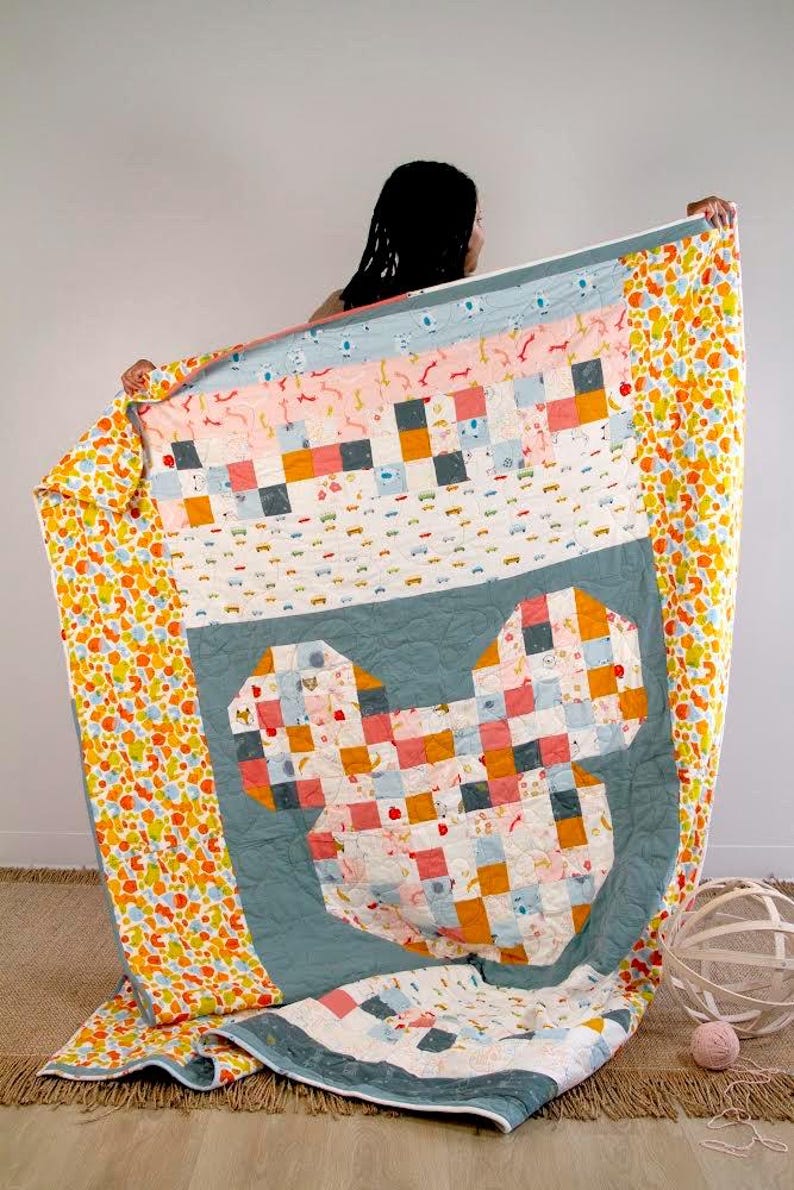 May include: A patchwork quilt with a large, heart-shaped design in the center. The quilt is made of various fabrics in shades of pink, blue, yellow, and orange. The quilt is folded and displayed on a wooden floor.