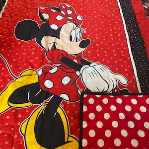 May include: A red and white polka dot quilt featuring a large Minnie Mouse character. The quilt has a red background with white polka dots and a black border with white polka dots. The Minnie Mouse character is wearing a red dress with white polka dots and a matching bow.