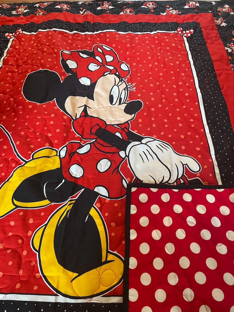 May include: A red and white polka dot quilt featuring a large Minnie Mouse character. The quilt has a red background with white polka dots and a black border with white polka dots. The Minnie Mouse character is wearing a red dress with white polka dots and a matching bow.