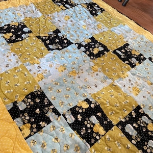 May include: A patchwork quilt with a yellow border and backing. The quilt top features squares of light blue, black, and yellow fabric, each patterned with bees and honeycombs. The quilt is machine-stitched with a wavy pattern.