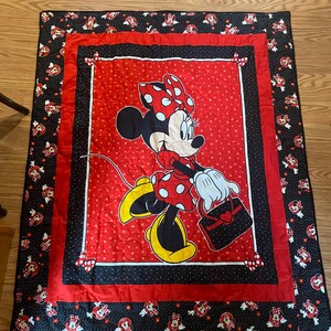 May include: A red and black Minnie Mouse quilt. The quilt features a large image of Minnie Mouse in a red polka dot dress and bow. The border is black with Minnie Mouse images and red accents. The quilt is likely for a child's bed.
