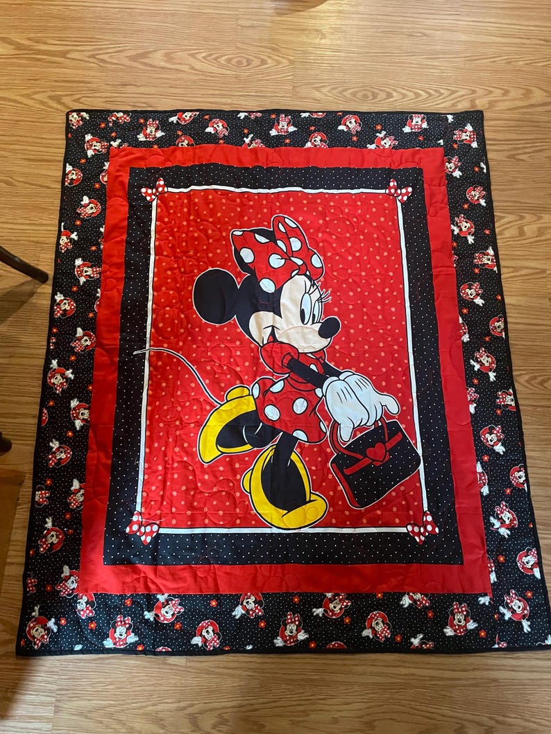 May include: A red and black Minnie Mouse quilt. The quilt features a large image of Minnie Mouse in a red polka dot dress and bow. The border is black with Minnie Mouse images and red accents. The quilt is likely for a child's bed.