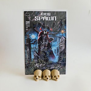 May include: A comic book titled "King Spawn" featuring a dark illustration of a cloaked figure in a graveyard setting. Three small, beige skulls are positioned in front of the comic book.