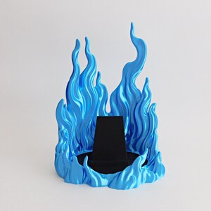 May include: A display stand featuring a bright blue, flame-shaped surround with a glossy finish. The center of the base contains a black, trapezoidal velvet-covered platform designed to hold items, creating a stark contrast with the blue flames.