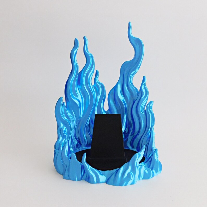 May include: A display stand featuring a bright blue, flame-shaped surround with a glossy finish. The center of the base contains a black, trapezoidal velvet-covered platform designed to hold items, creating a stark contrast with the blue flames.