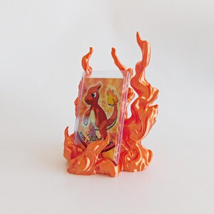 May include: A trading card featuring a red lizard character is displayed in a clear plastic protective sleeve, which is held upright by a decorative orange and yellow flame-shaped stand with sculpted, swirling fire details.