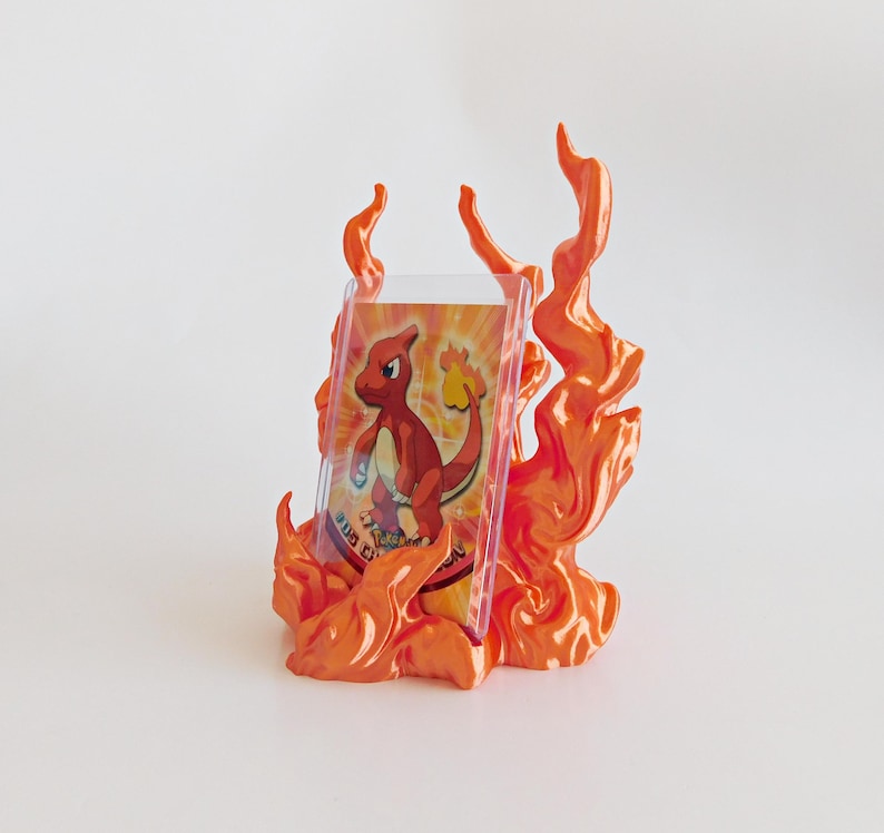May include: A trading card featuring a red lizard character is displayed in a clear plastic protective sleeve, which is held upright by a decorative orange and yellow flame-shaped stand with sculpted, swirling fire details.