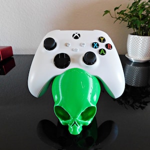 May include: A white Xbox controller is resting on a green alien skull-shaped stand. The controller has black buttons and a green Xbox logo.
