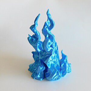May include: A bright blue, glossy, three-dimensional sculpture shaped like stylized, upward-reaching flames. The object features a smooth, reflective surface with intricate, swirling curves and pointed peaks that mimic the appearance of fire.