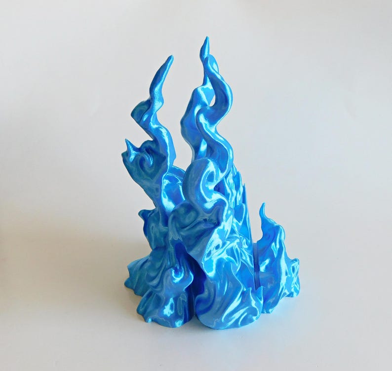 May include: A bright blue, glossy, three-dimensional sculpture shaped like stylized, upward-reaching flames. The object features a smooth, reflective surface with intricate, swirling curves and pointed peaks that mimic the appearance of fire.