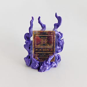 May include: A rectangular trading card featuring a gold-colored Mew character illustration and ancient-style symbols, encased in a clear protective sleeve and mounted within a three-dimensional, glossy purple flame-shaped display stand.