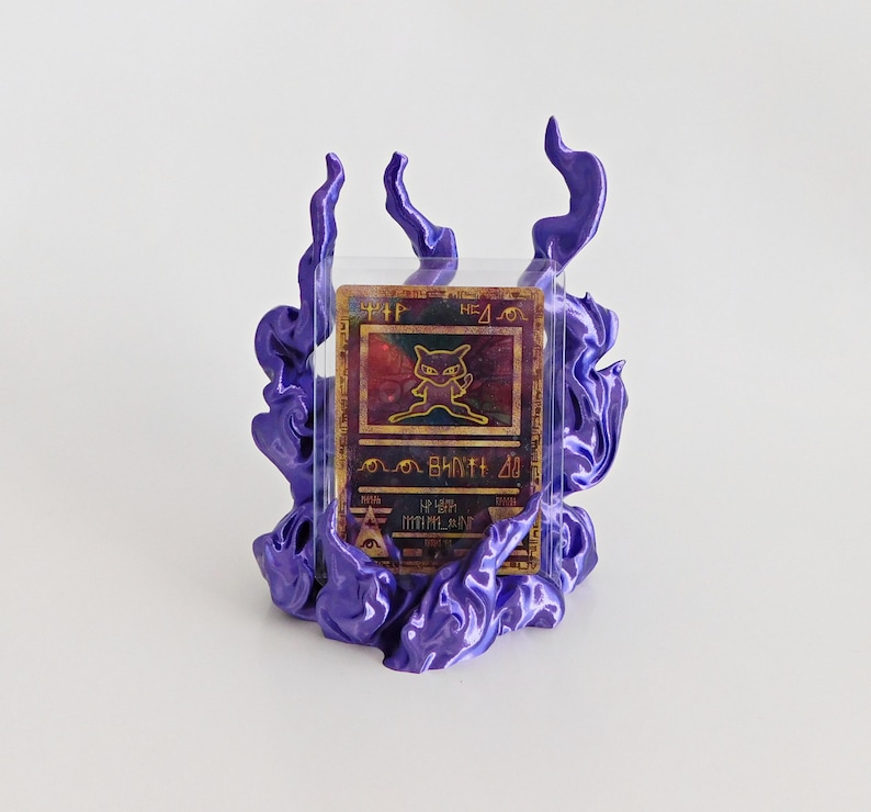 May include: A rectangular trading card featuring a gold-colored Mew character illustration and ancient-style symbols, encased in a clear protective sleeve and mounted within a three-dimensional, glossy purple flame-shaped display stand.