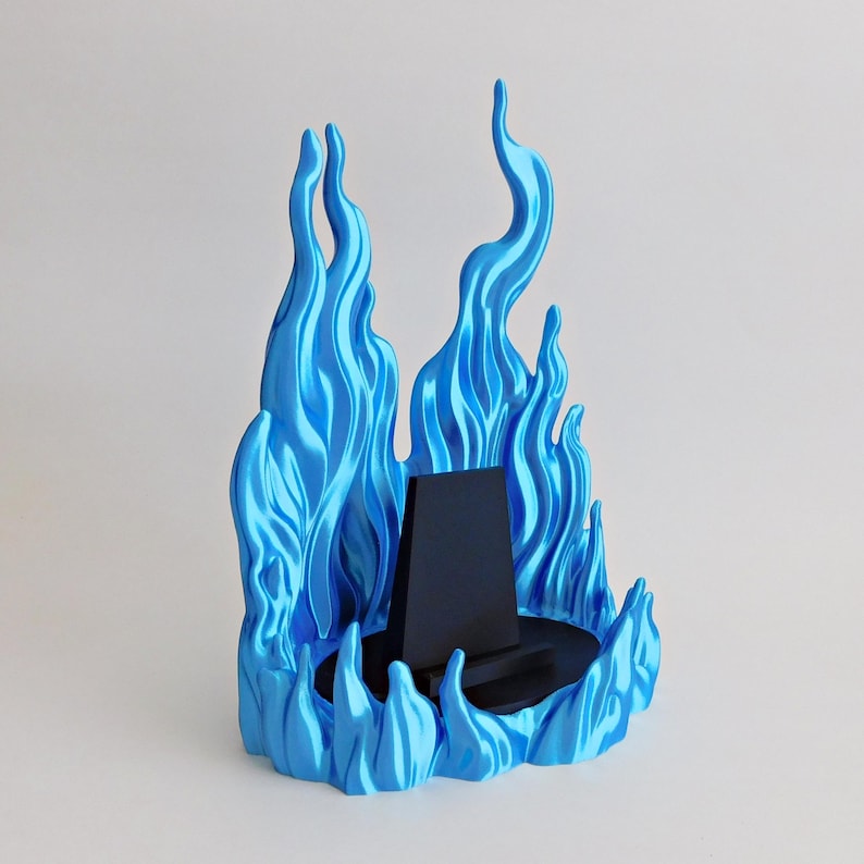 May include: A display stand featuring a central black trapezoidal support mounted on a circular black base, surrounded by a sculpted, metallic blue flame-like structure that rises vertically to create a decorative border around the center.