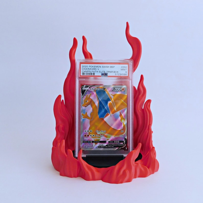 May include: A graded 2020 Pokemon Charizard V trading card in a clear plastic PSA slab, displayed upright in a bright red, three-dimensional flame-shaped stand. The card features a holographic Charizard illustration with 220 HP and text.