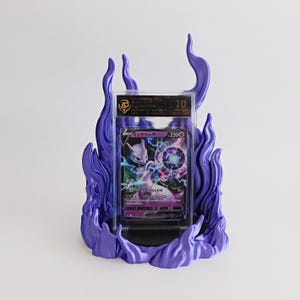 May include: A graded Mewtwo V trading card encased in a clear plastic slab, displayed within a purple flame-shaped stand. The card features a purple and black color scheme with Japanese text and a 220 HP value. The slab displays a grade of 10.