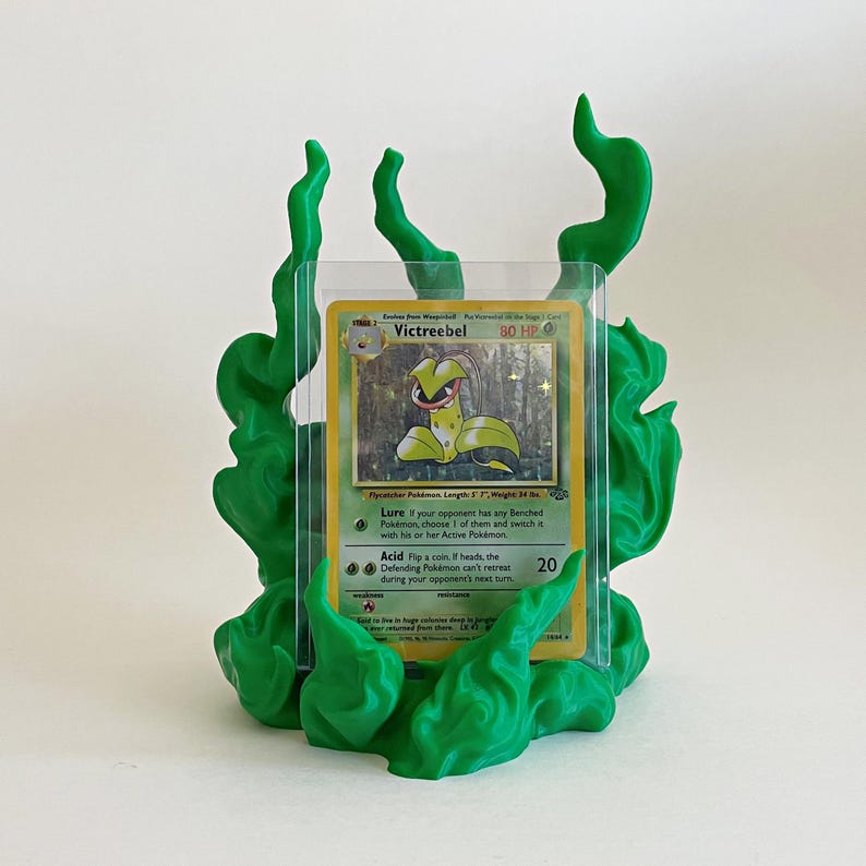 May include: A green plastic display stand shaped like stylized flames holding a Victreebel trading card in a clear protective sleeve. The card features the character Victreebel with 80 HP, listing Lure and Acid attacks, and text detailing its physical traits.
