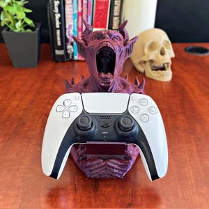 May include: A burgundy-colored demon-themed controller holder with an open mouth design. The holder supports a white and black PlayStation 5 controller. A skull and books are in the background.