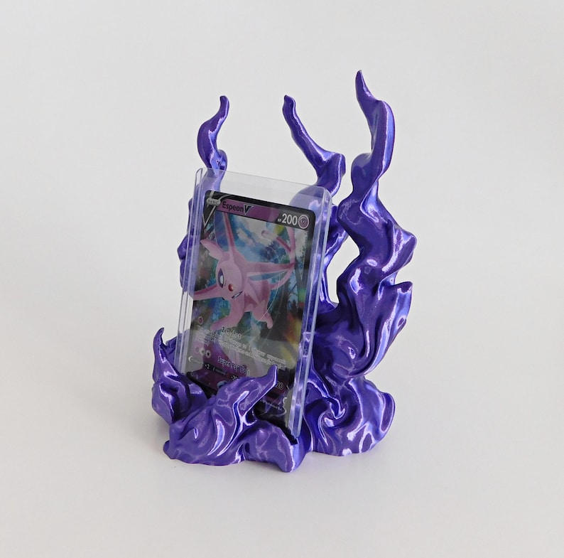 May include: A purple display stand shaped like stylized flames holding a clear plastic protective case containing a pink Espeon V trading card with 200 HP. The stand features a glossy finish and organic, swirling flame-like structures.