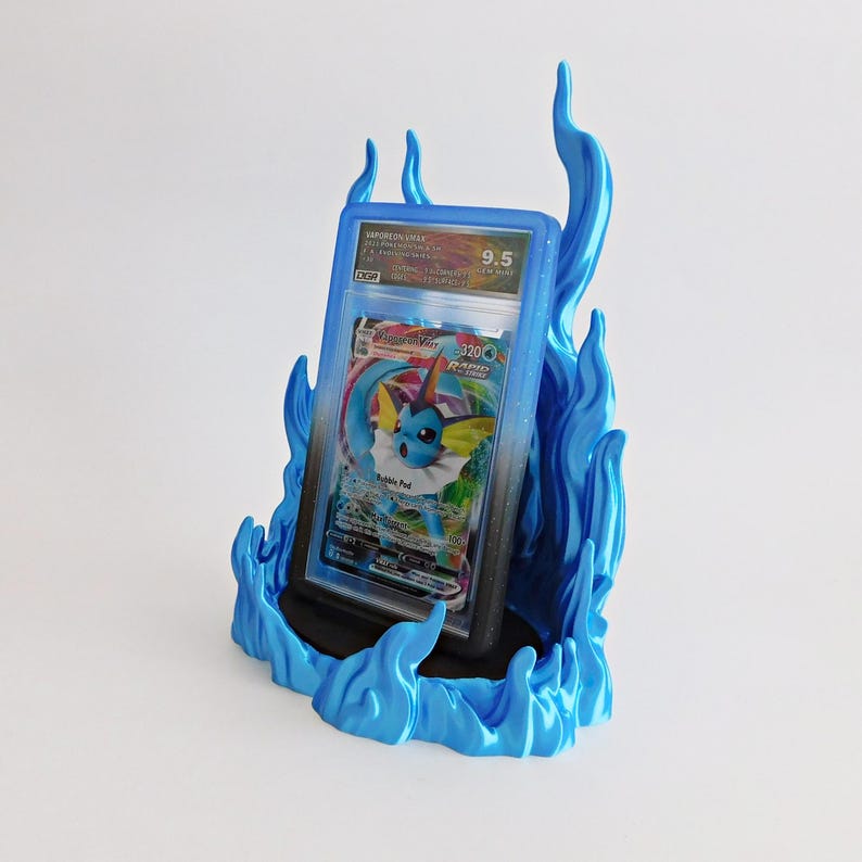 May include: A graded Vaporeon VMAX trading card encased in a clear plastic slab, displayed within a bright blue, flame-shaped stand. The card features a blue creature with yellow ears and a white collar, with the text Vaporeon VMAX and 320 HP visible.