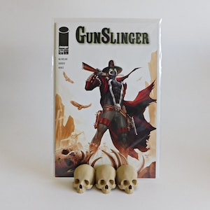 May include: A comic book titled "Gunslinger" featuring a character in a black and red costume with a gun. Three small, beige skulls are in front of the comic book.