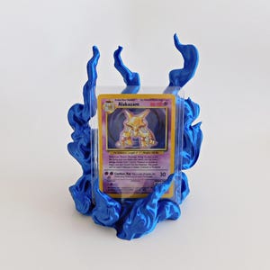 May include: A blue plastic display stand shaped like stylized flames holding a clear protective sleeve containing a Pok&eacute;mon trading card. The card features the character Alakazam with yellow and purple colors and printed text including the name Alakazam.