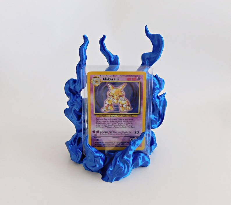 May include: A blue plastic display stand shaped like stylized flames holding a clear protective sleeve containing a Pok&eacute;mon trading card. The card features the character Alakazam with yellow and purple colors and printed text including the name Alakazam.