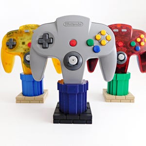 May include: Three Nintendo 64 game controllers displayed on stands. The center controller is solid gray, the left is translucent yellow, and the right is translucent red. Each controller rests on a vertical pipe-shaped base with a brick-style foundation.