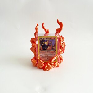 May include: A Dark Charizard trading card encased in a clear protective plastic sleeve, positioned within a decorative orange flame-shaped stand. The card features a dragon illustration and text including Dark Charizard, Nail Flick, and Continuous Fireball.