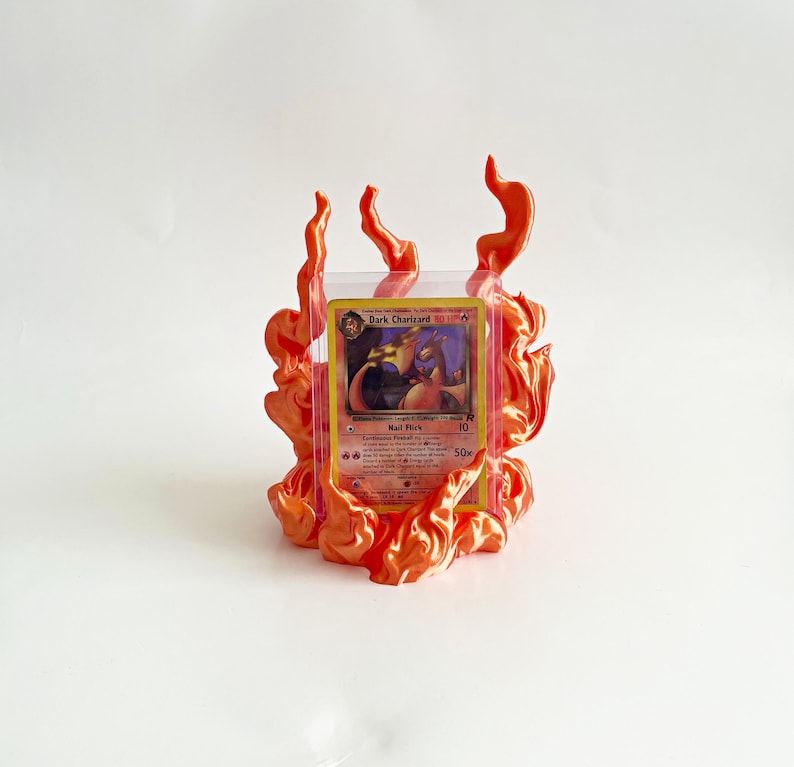 May include: A Dark Charizard trading card encased in a clear protective plastic sleeve, positioned within a decorative orange flame-shaped stand. The card features a dragon illustration and text including Dark Charizard, Nail Flick, and Continuous Fireball.