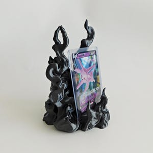 May include: A black, flame-shaped display stand holding a clear plastic protective case containing a purple Espeon V trading card. The stand features swirling, smoke-like contours that rise upward to support the card in an upright position.