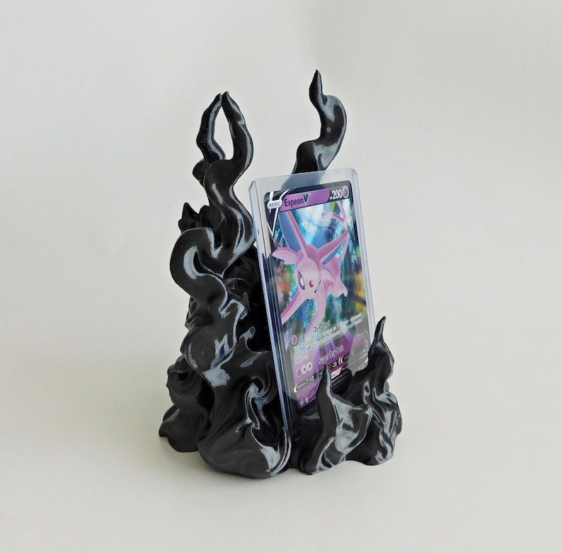 May include: A black, flame-shaped display stand holding a clear plastic protective case containing a purple Espeon V trading card. The stand features swirling, smoke-like contours that rise upward to support the card in an upright position.