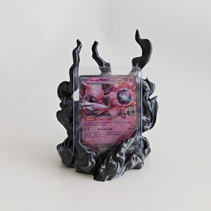 May include: A black display stand featuring sculpted, flame-like smoke patterns holding a clear plastic protective sleeve. Inside the sleeve is a Team Rocket's Mewtwo GX trading card with pink and purple artwork, displaying 280 HP and text.