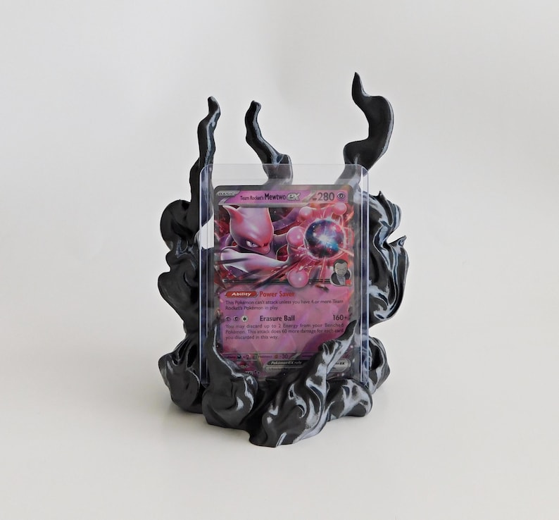 May include: A black display stand featuring sculpted, flame-like smoke patterns holding a clear plastic protective sleeve. Inside the sleeve is a Team Rocket's Mewtwo GX trading card with pink and purple artwork, displaying 280 HP and text.