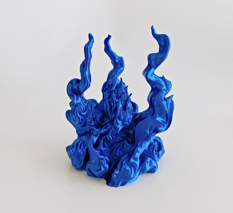 May include: A metallic blue figurine depicting a central figure surrounded by swirling, flame-like structures. The object features a glossy, reflective finish with intricate, sculpted details that mimic the appearance of rising smoke or fire.