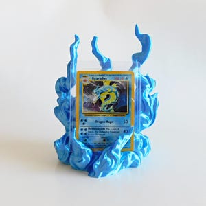 May include: A trading card display stand featuring a sculpted blue flame design surrounding a clear plastic sleeve. The card inside depicts a yellow and blue Gyarados character with the text Gyarados, Dragon Rage, and Bubblebeam printed on it.