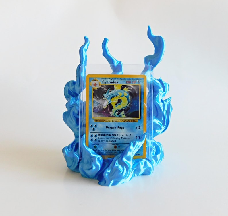 May include: A trading card display stand featuring a sculpted blue flame design surrounding a clear plastic sleeve. The card inside depicts a yellow and blue Gyarados character with the text Gyarados, Dragon Rage, and Bubblebeam printed on it.