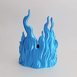 May include: A bright blue decorative object shaped like stylized flames with a smooth, glossy finish. The structure features multiple vertical, undulating peaks of varying heights that create a continuous, wave-like pattern around the perimeter.