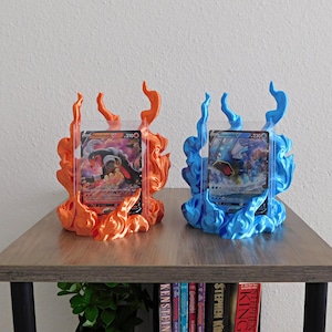 May include: Two 3D-printed display stands for trading cards, one in glossy orange flame shapes and one in glossy blue flame shapes. Each stand holds a clear plastic card protector containing a Houndoom V card and a Gyarados V card respectively.