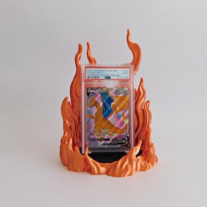 May include: A graded Charizard V trading card encased in a clear plastic slab, positioned within an orange flame-shaped display stand. The card label reads 2020 POKEMON SWSH BSP CHARIZARD V CHMPN PATH ELITE TRNR BOX #050 MINT 9 57234588.
