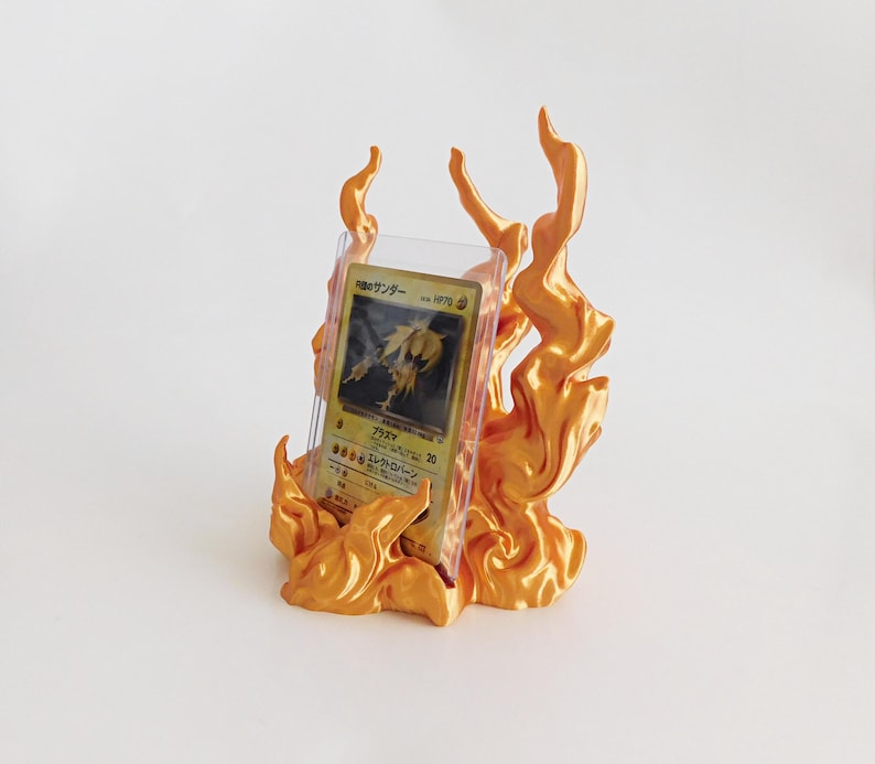 May include: A metallic orange display stand shaped like stylized flames, holding a Japanese trading card encased in a clear plastic protective sleeve. The card features a yellow creature illustration and Japanese text including HP70.