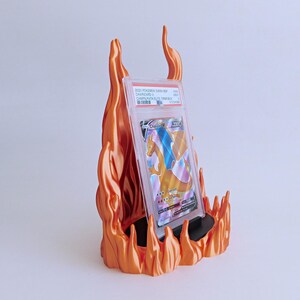 May include: A graded Charizard V trading card in a clear plastic protective case, displayed on a black stand surrounded by a sculpted orange flame-shaped holder. The card label reads 2020 POKEMON SWSH BSP CHARIZARD V CHMPN PATH ELITE TRNR BOX.