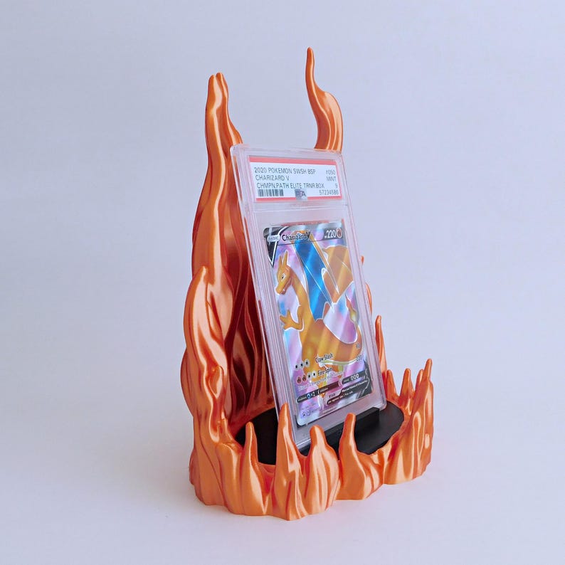 May include: A graded Charizard V trading card in a clear plastic protective case, displayed on a black stand surrounded by a sculpted orange flame-shaped holder. The card label reads 2020 POKEMON SWSH BSP CHARIZARD V CHMPN PATH ELITE TRNR BOX.