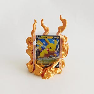 May include: A Pikachu V trading card encased in a clear plastic protective sleeve, positioned within a three-dimensional, metallic gold-colored flame-shaped display stand that surrounds the card on the sides and bottom.
