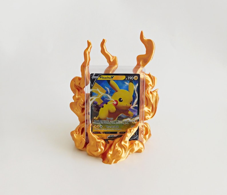 May include: A Pikachu V trading card encased in a clear plastic protective sleeve, positioned within a three-dimensional, metallic gold-colored flame-shaped display stand that surrounds the card on the sides and bottom.